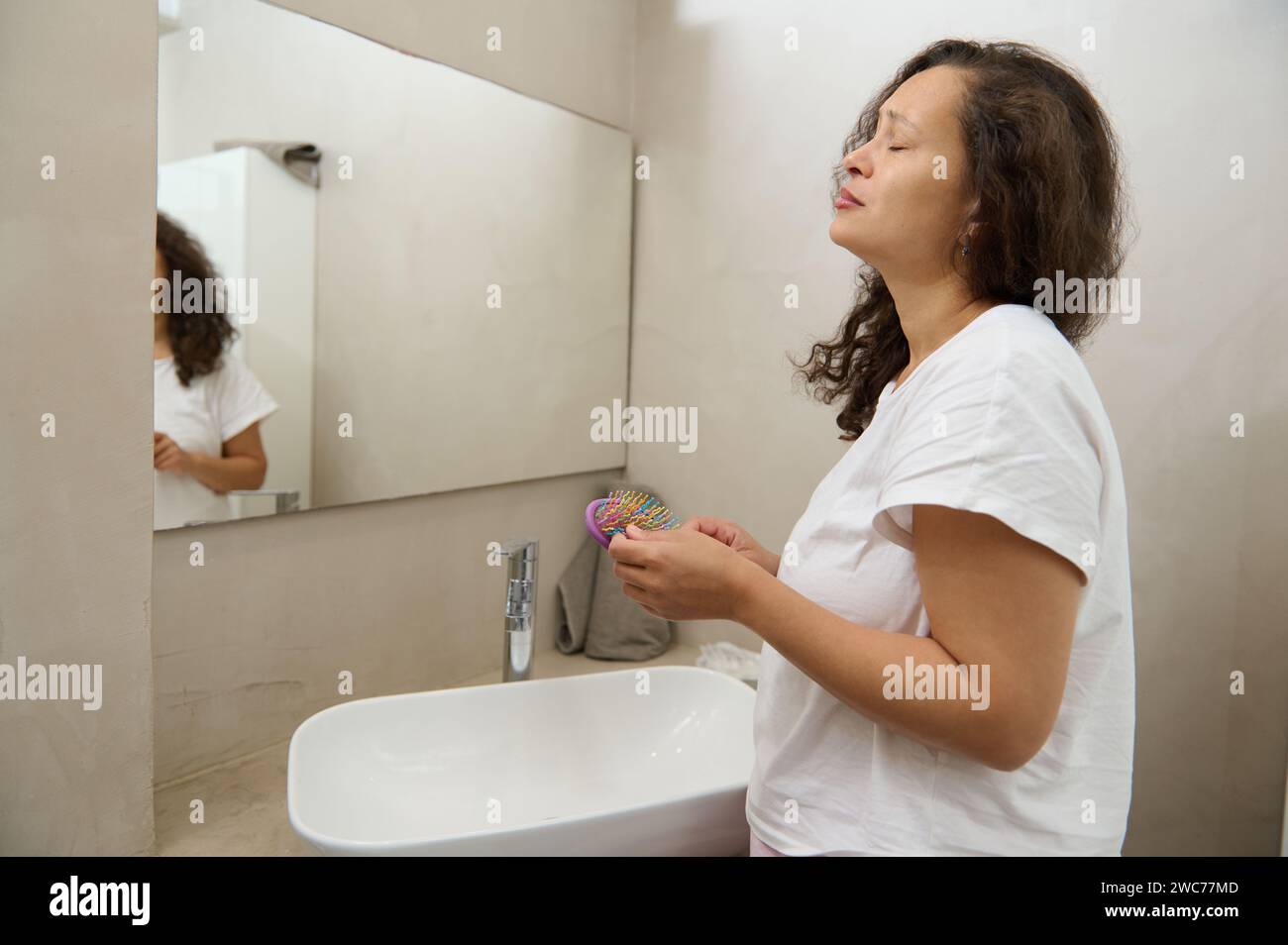 Besorgte junge Frau reflektiert sich im Spiegel, besorgt über Haarausfall, kämmt im Spiegel, hält Haarbürste und Sperre. Verärgerte junge Frau, die fe zählt Stockfoto