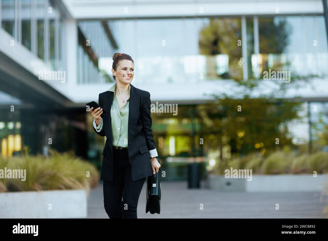 Glückliche moderne 40-jährige Arbeiterin in der Nähe des Business Centers in schwarzer Jacke mit Aktentasche mit Smartphone und zu Fuß. Stockfoto