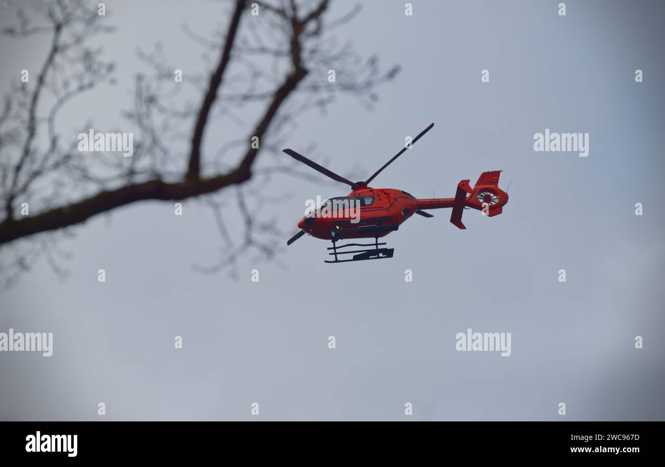 Orange-roter Hubschrauber des Rettungsdienstes am hellgrauen Himmel und ein dunkler Ast im Vordergrund (außer Fokus) Stockfoto