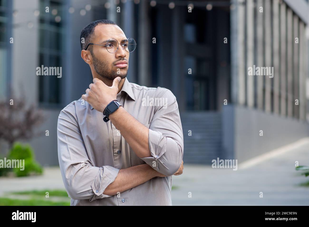 Nachdenklicher junger Mann mit Brille und Smartwatch in urbaner Umgebung, Hand am Kinn, besinnend, mit einem fokussierten Ausdruck. Stockfoto