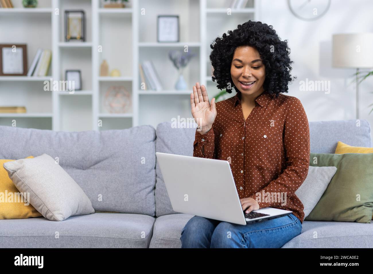 Lächelnde afroamerikanische junge Frau sitzt zu Hause auf dem Sofa, spricht per Laptop per Videoanruf, winkt und grüßt vor der Kamera. Stockfoto