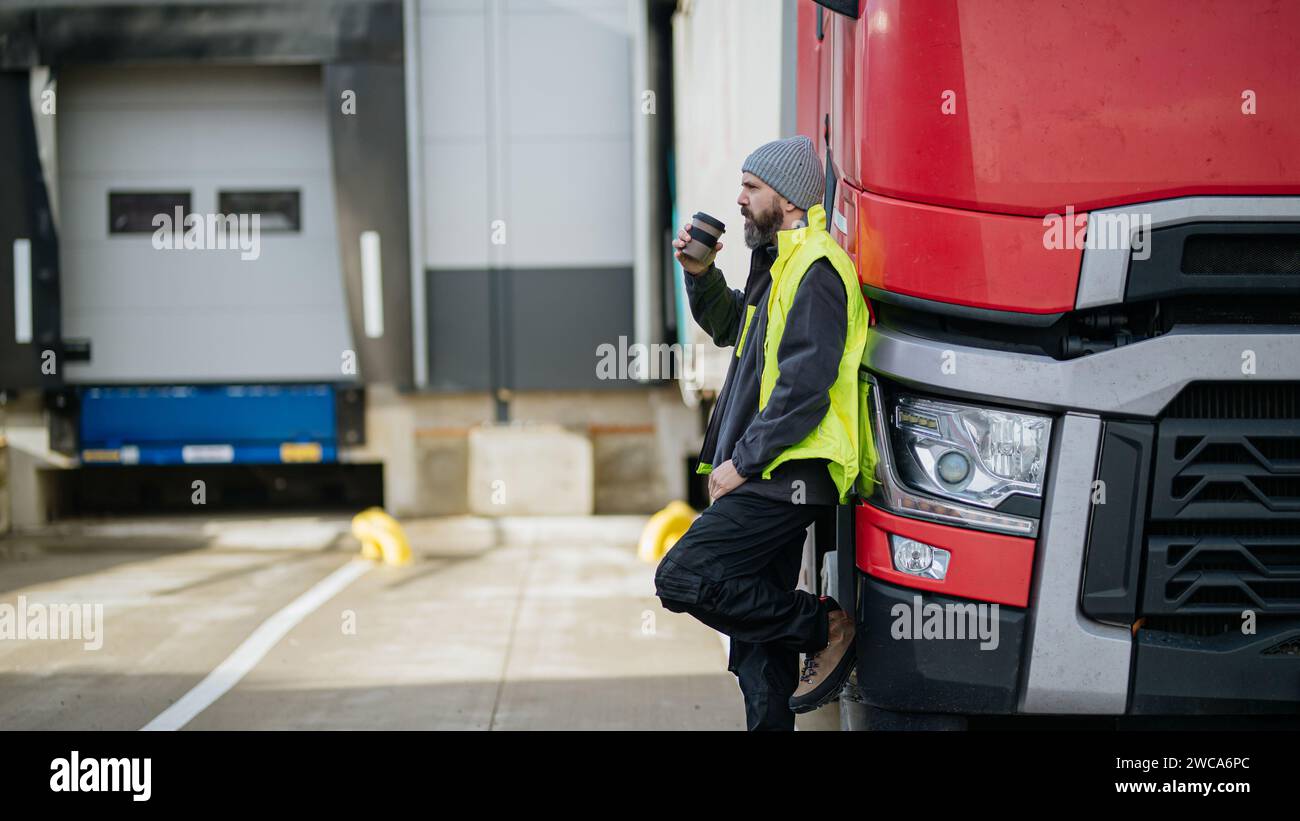 Lkw-Fahrer lehnt sich an den roten Lkw und trinkt Kaffee, wartet auf Lagerarbeiter. Stockfoto