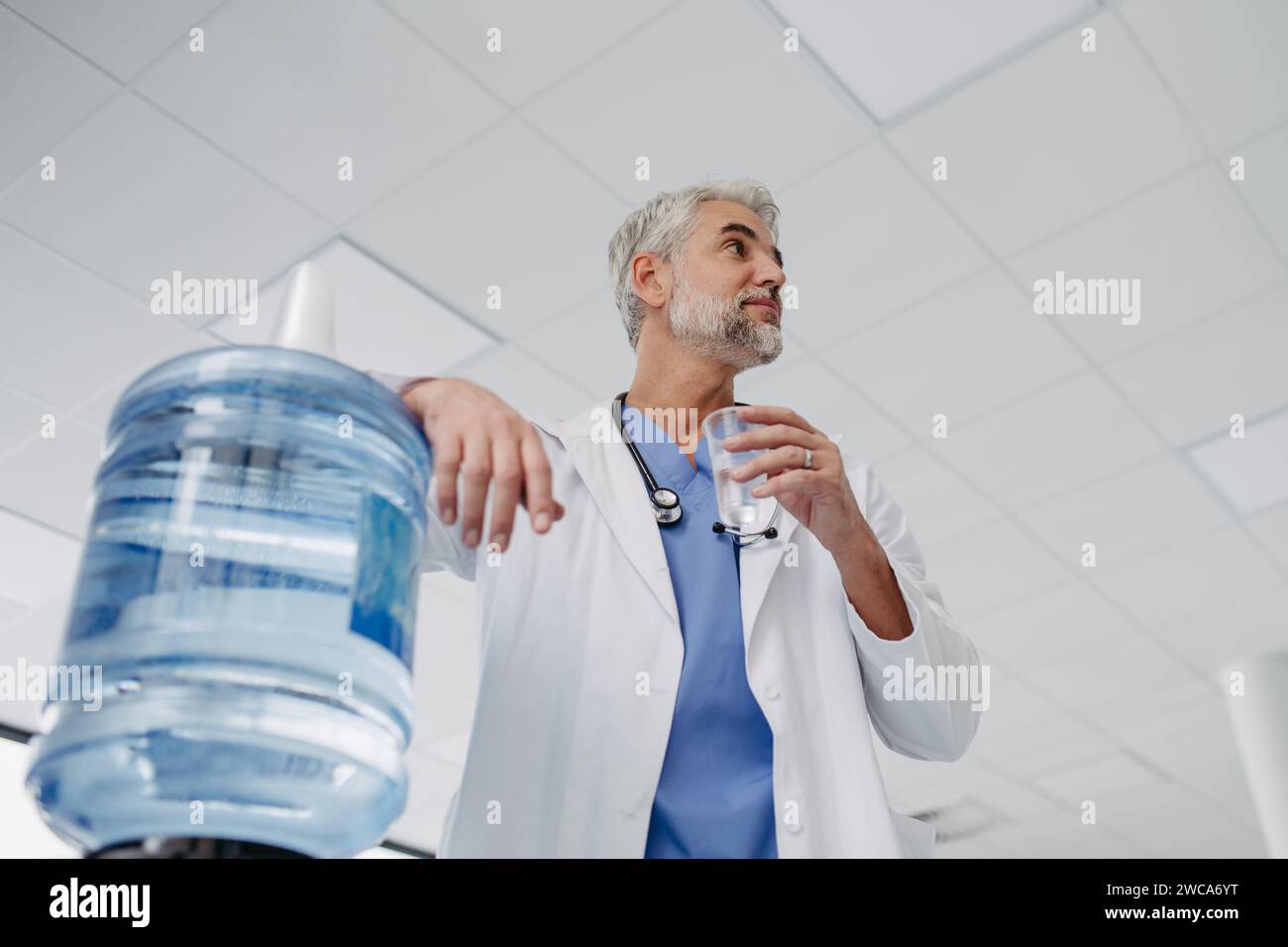 Gutaussehender Arzt, der eine Pause während der Arbeitsschicht im Krankenhaus macht, Trinkwasser aus dem Wasserspender in der Krankenhauslobby trinkt. Stockfoto