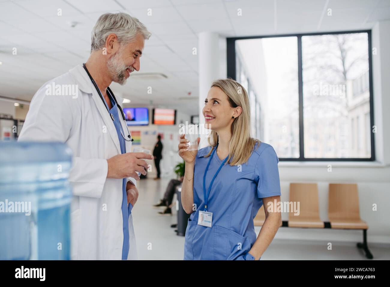 Arzt und Krankenschwester machen eine Pause während der Arbeitsschicht im Krankenhaus, trinken Wasser aus dem Wasserspender in der Krankenhauslobby, sprechen. Stockfoto