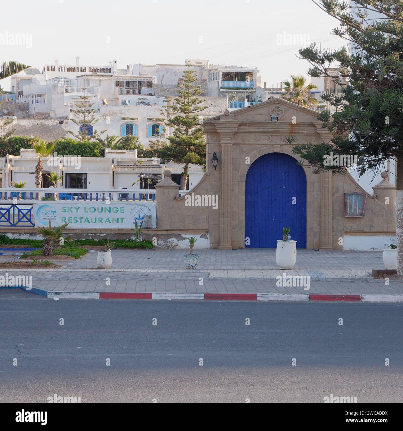 Straße mit roten und weißen Bordsteinen und einem kunstvollen Eingangstor mit blauer Holztür in Essaouira Marokko, 15. Januar 2024 Stockfoto