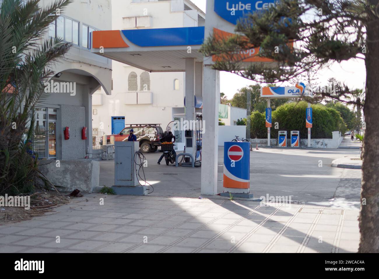 Blau-orange Tankstelle mit einem Mann auf einem Motorrad mit Orangen in einer Kiste, in Essaouira Marokko, 14. Januar 2024 Stockfoto