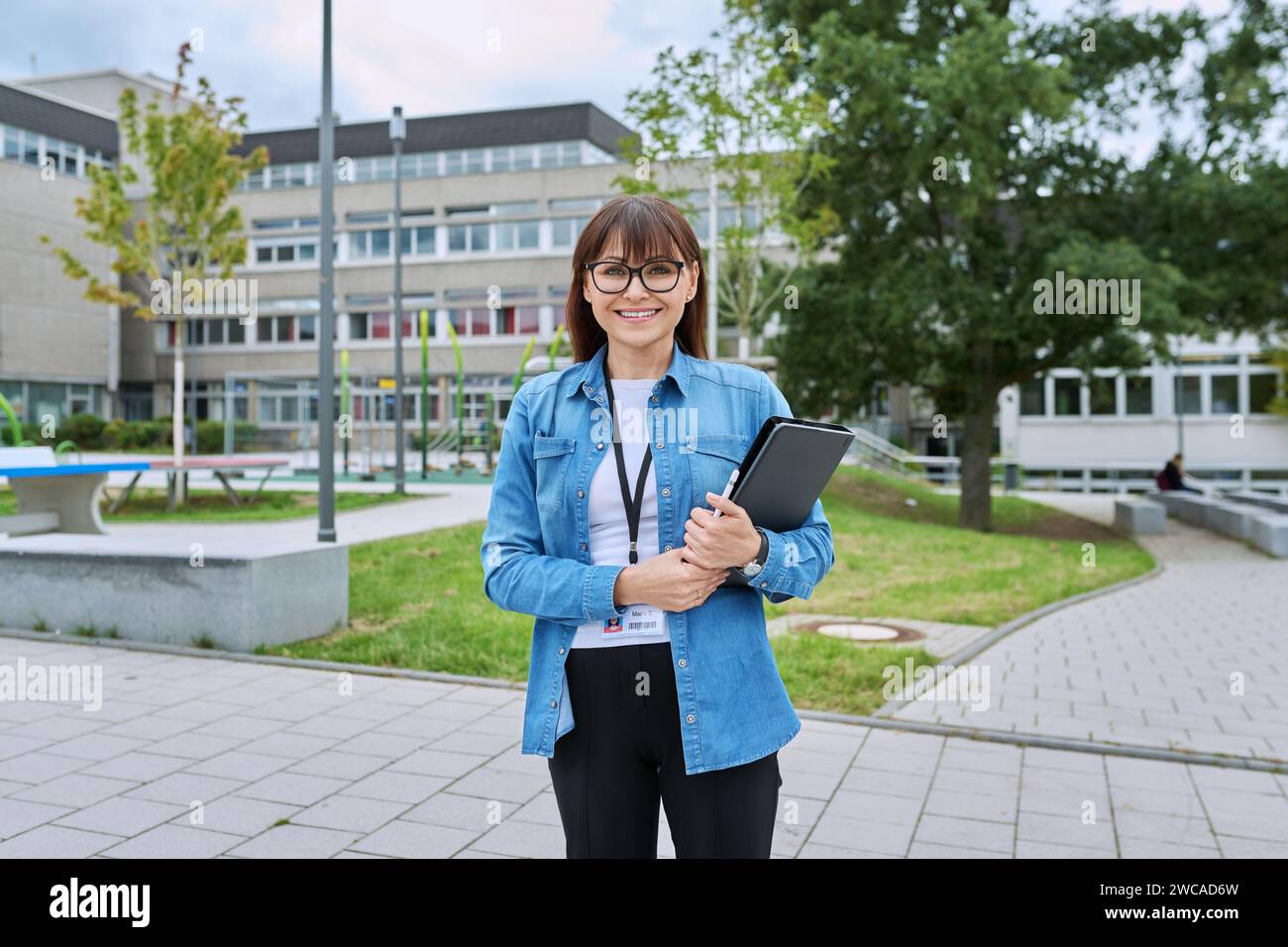 Schullehrerin mittleren Alters mit digitalem Tablet posiert in der Nähe des Schulgebäudes Stockfoto