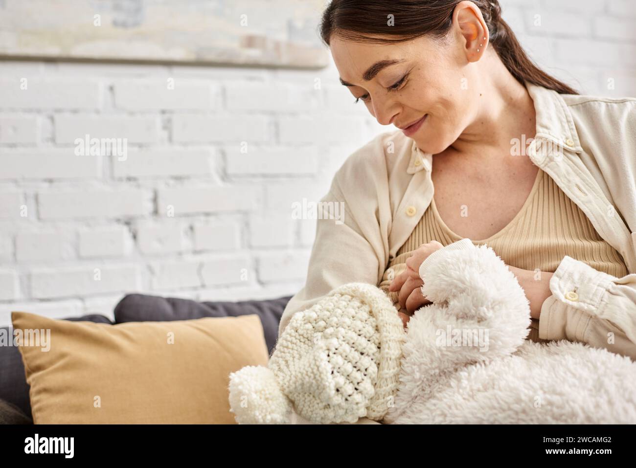 Attraktive fürsorgliche, fröhliche Mutter in kuscheliger Hauswäsche, die ihr neugeborenes Kind stillt, moderne Erziehung Stockfoto