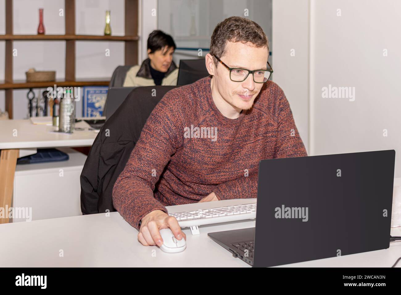Ein Sekretär, der mit seinem Laptop im Büro arbeitet, während sein Kollege im Hintergrund an einem anderen Schreibtisch arbeitet Stockfoto