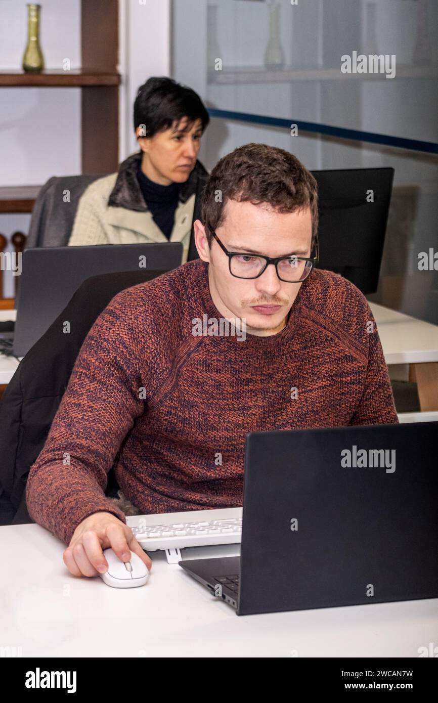 Ein Sekretär, der mit seinem Laptop im Büro arbeitet, während sein Kollege im Hintergrund an einem anderen Schreibtisch arbeitet Stockfoto