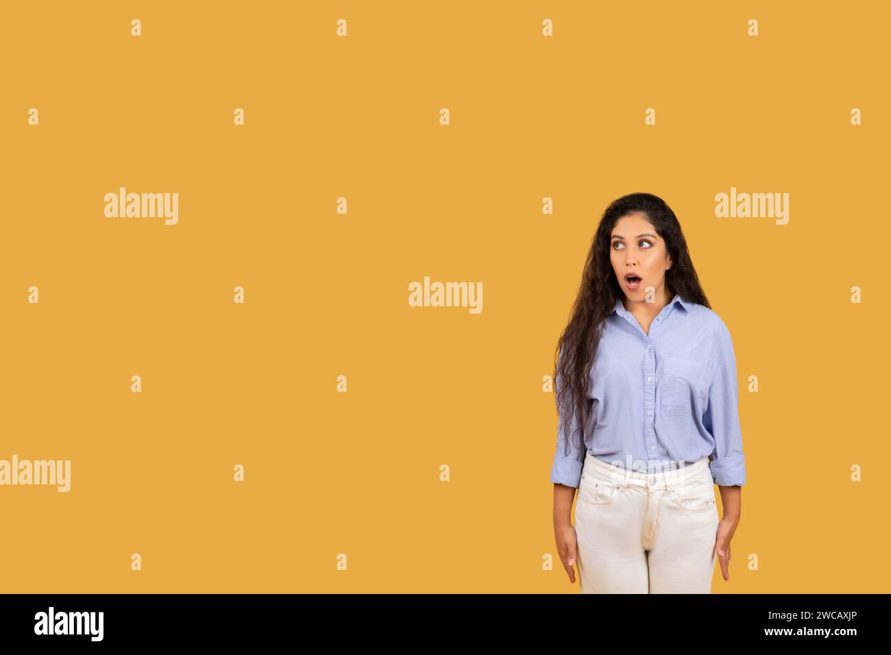 Überraschte junge Frau mit langen, welligen Haaren und breiten Augen, die ein blaues Hemd trägt Stockfoto