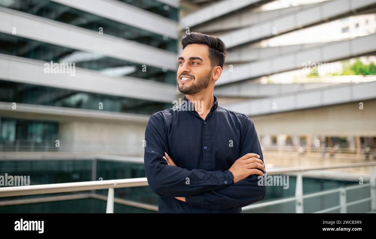 Ein lächelnder junger Geschäftsmann aus dem Nahen Osten steht im Freien selbstbewusst über seine Arme Stockfoto