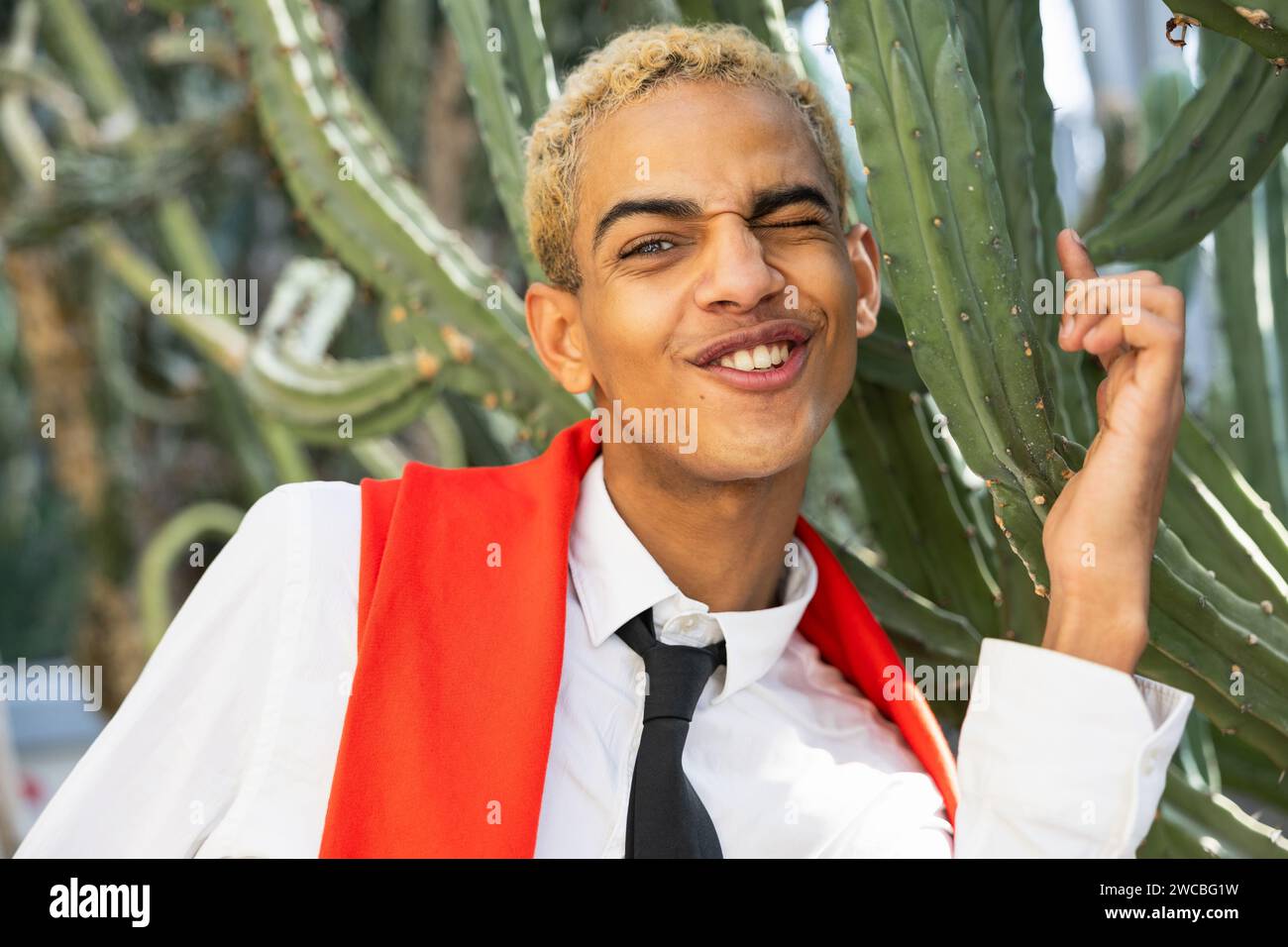 Ein fröhlicher junger Mann zwinkert und streckt seine Zunge aus, während er verspielt vor einem großen Kakteen in einem botanischen Garten posiert. Stockfoto