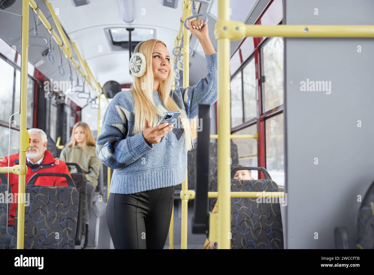 Seitenansicht eines stilvollen blonden Mädchens mit Telefon, das auf das lächelnde Fenster blickt, das in sich bewegenden öffentlichen Verkehrsmitteln steht. Besatz des weiblichen Haltegriffs mit verschwommenen Fahrgästen im Bus. Konzept des Stadtlebens. Stockfoto