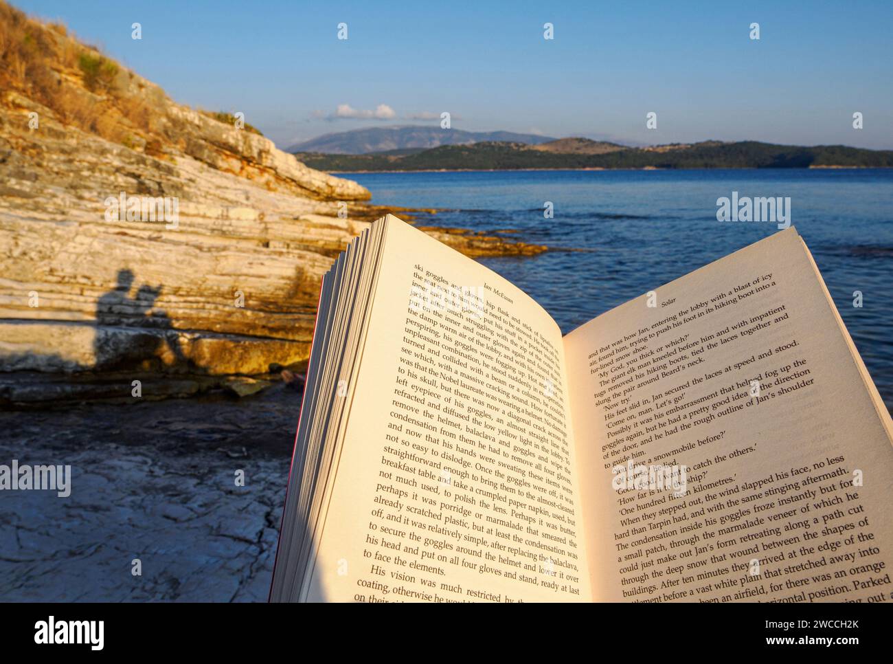 Mann liest Ian McEwans Roman Solar im Urlaub auf Korfu mit abendlicher Sonneneinstrahlung, die einen Schatten auf die Küstenklippen wirft - Ionische Inseln Griechenland Stockfoto