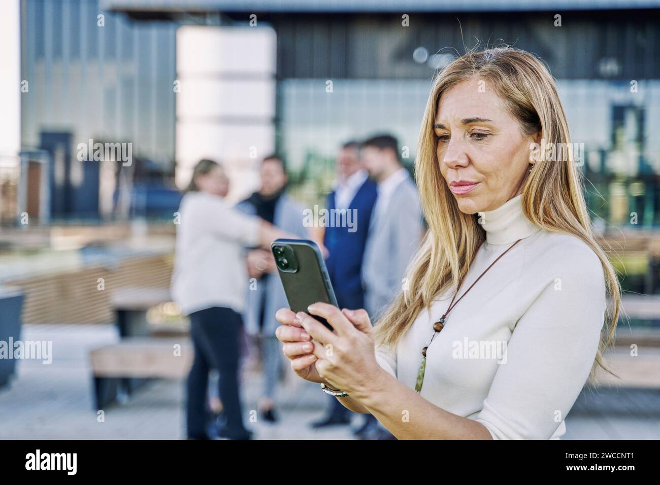 Eine 50-jährige Geschäftsfrau beherrscht ihr Mobilgerät im Vordergrund, während ihr engagiertes Team von Geschäftsleuten im Hintergrund harmonisch arbeitet Stockfoto