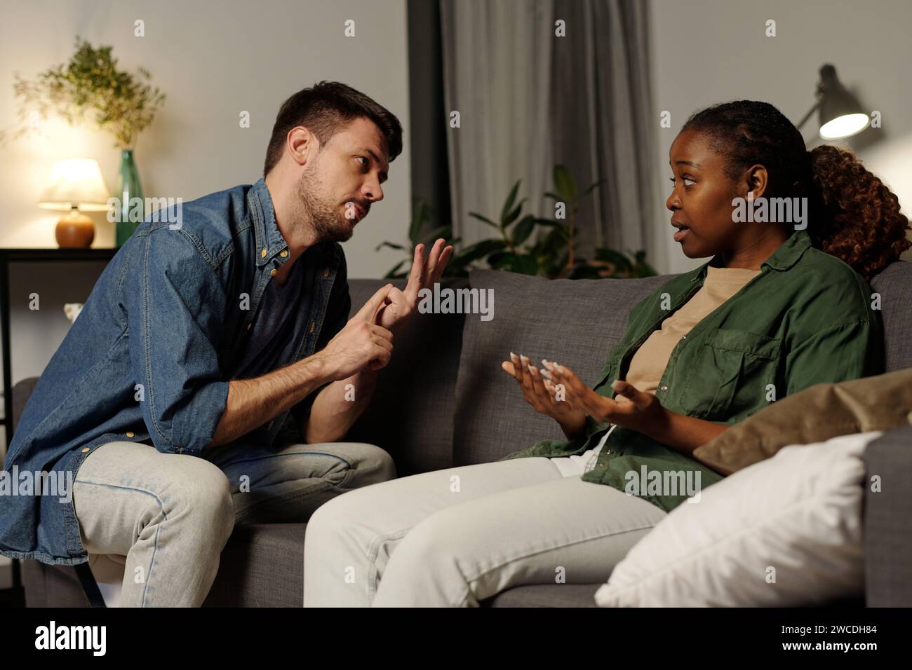 Eine junge irritierte Frau, die ihrem wütenden und eifersüchtigen Ehemann etwas erklärt, während sie vor ihm auf der Couch sitzt Stockfoto