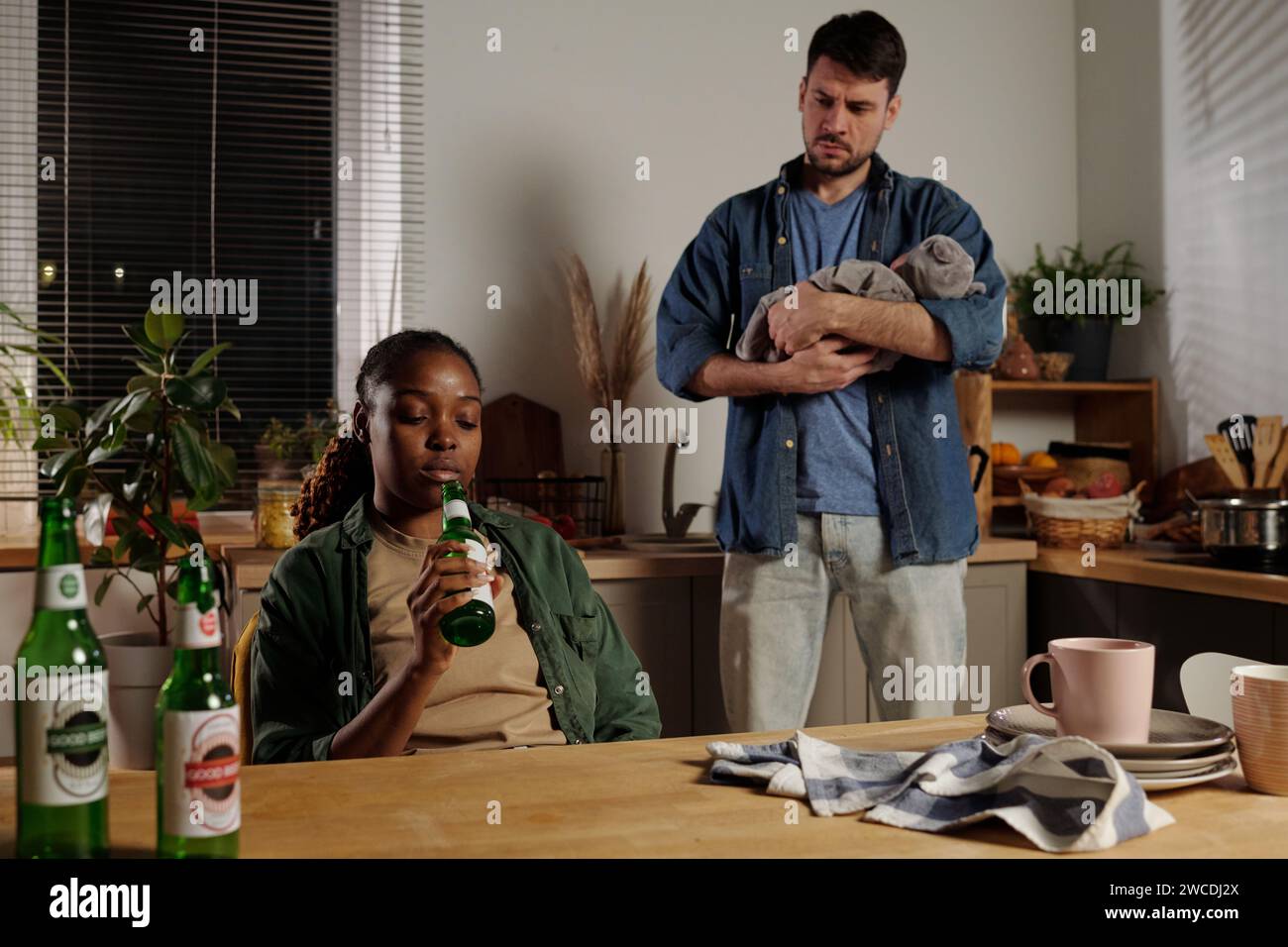 Junger Mann mit Baby auf den Händen, der auf seine verstörte Frau mit postnataler Depression blickt und am Tisch in der Küche sitzt und Bier trinkt Stockfoto