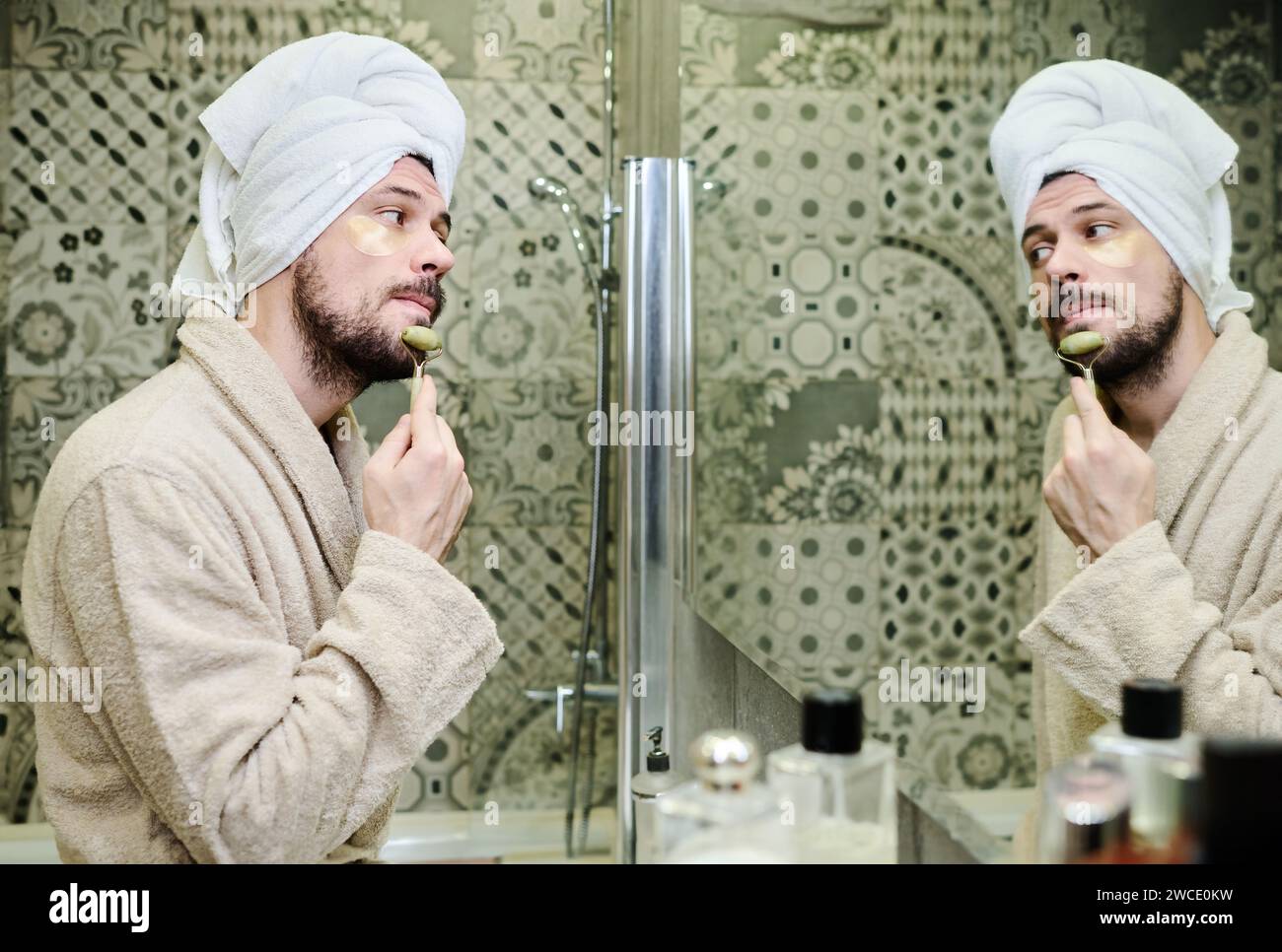 Junger Mann im Bademantel, der nach der Morgendusche vor dem Spiegel eine Gesichtsmassage mit Jadesteinrolle hat Stockfoto