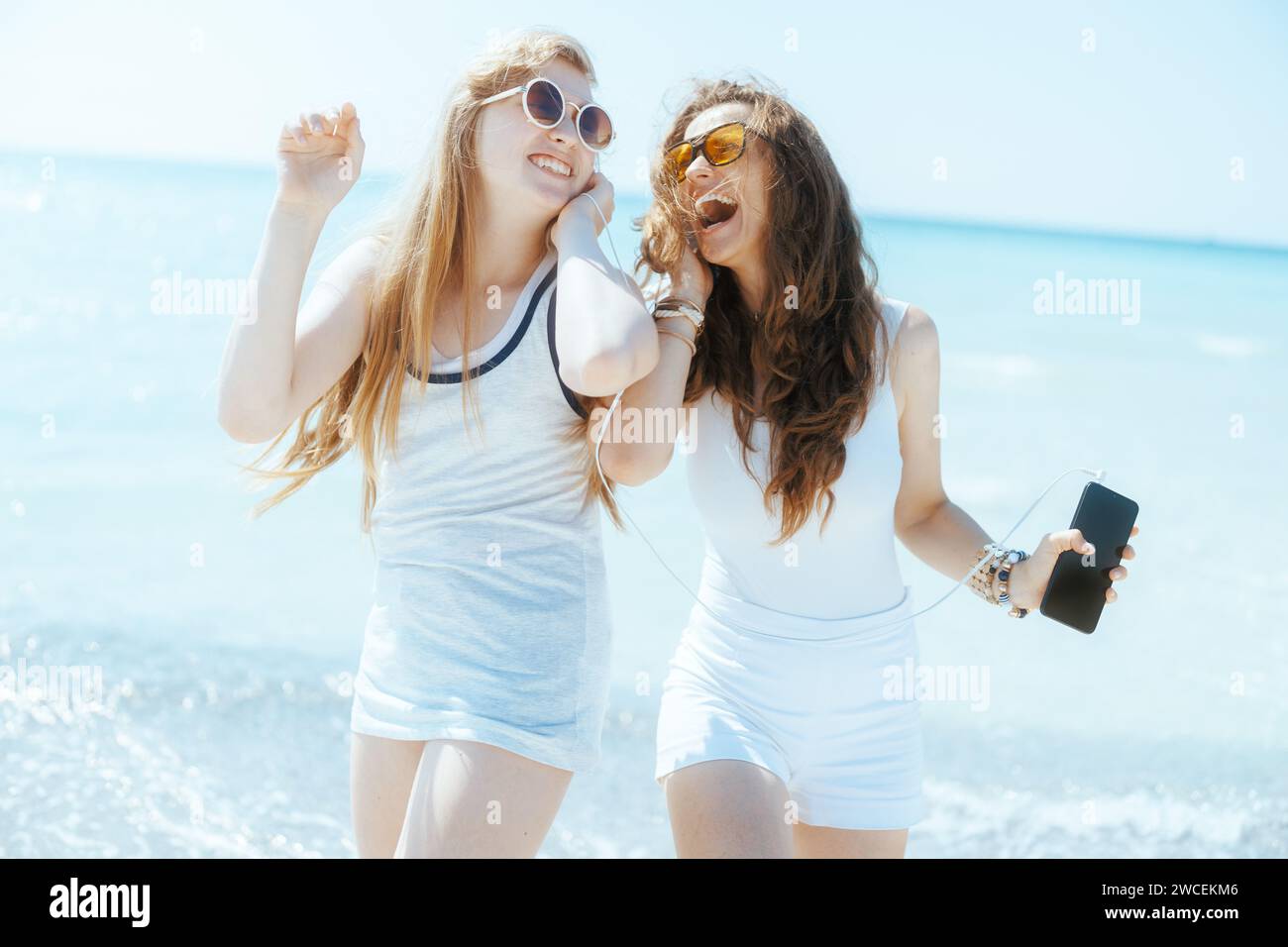 Lächelnde, moderne Mutter und Kind am Meer mit Smartphone, hören Musik mit Kopfhörern und tanzen. Stockfoto