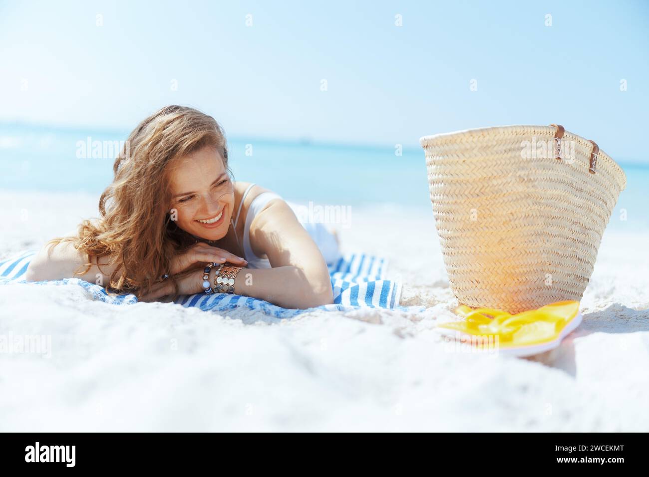 Glückliche, stilvolle Frau an der Meeresküste mit Strohbeutel und gestreiftem Handtuch. Stockfoto