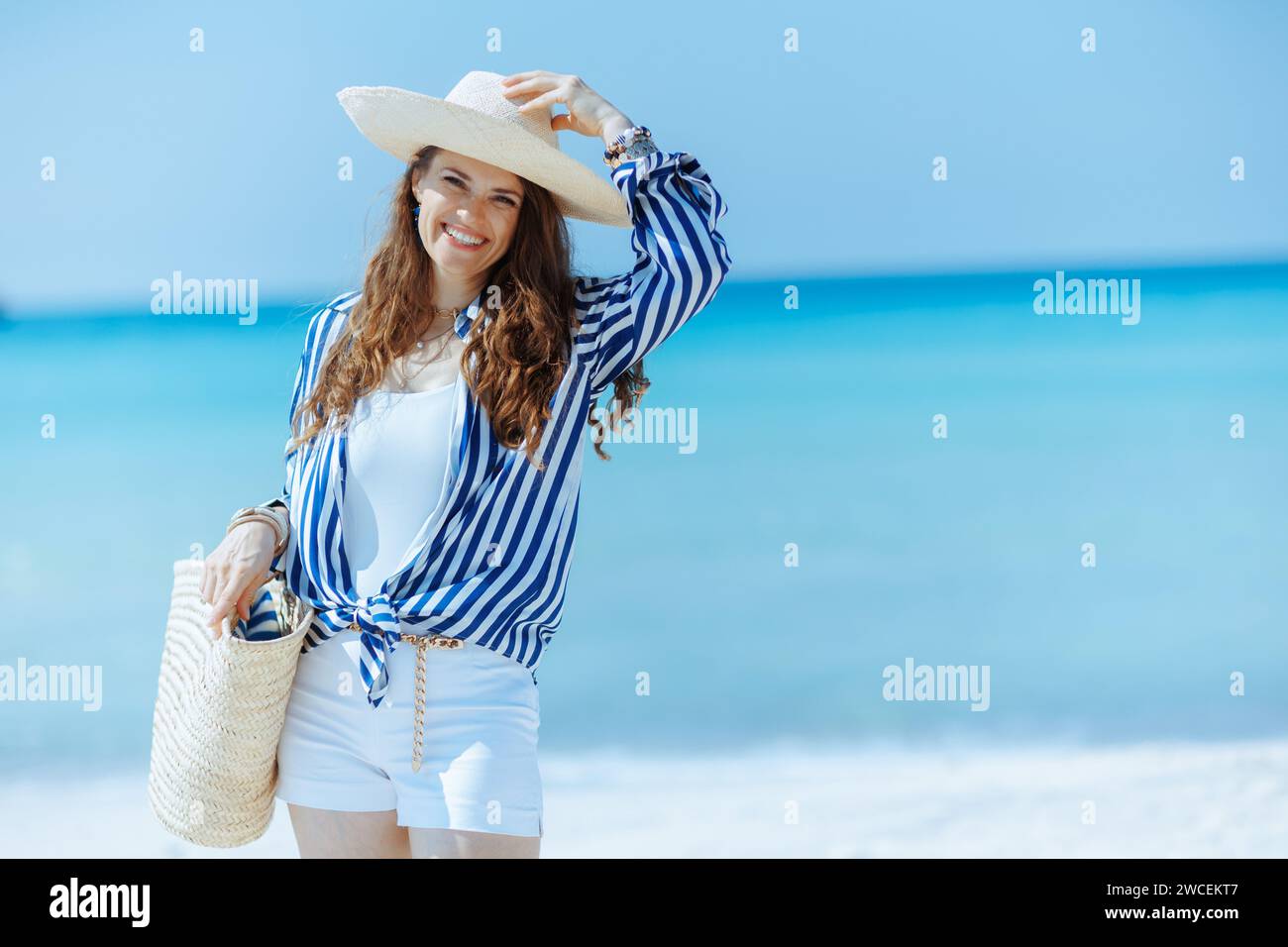 Lächelnde, elegante Frau am Meer mit Strohbeutel und Strohhut. Stockfoto