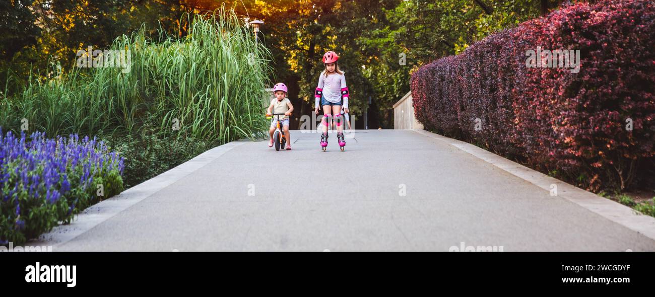 Zwei kleine Mädchen fahren auf Rollern und Laufrädern im Sommerpark. Kinder tragen Schutzpolster und Schutzhelm für eine sichere Fahrt. Aktiver Outdoor-Sport f Stockfoto