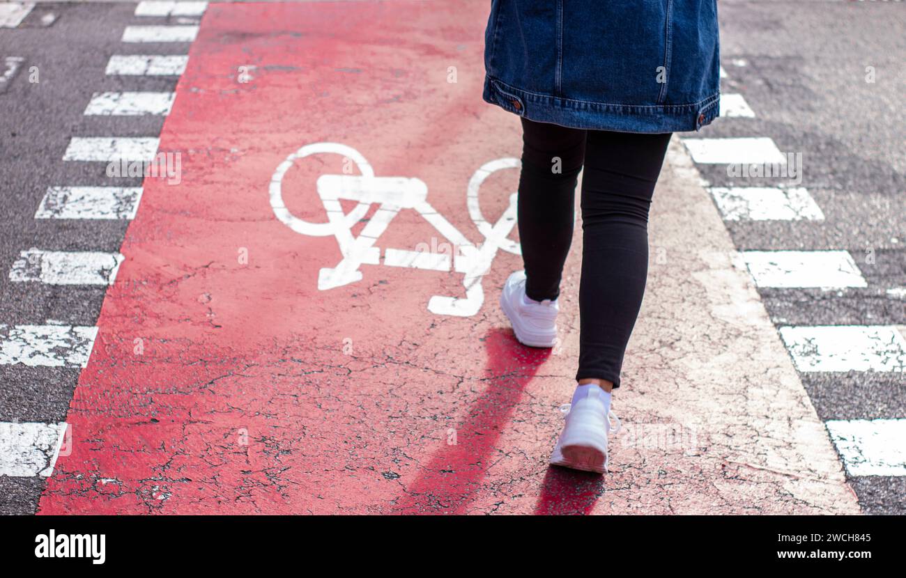 Das Mädchen in weißen Sneakers mit einer Jeansjacke geht zu Fuß über einen Fahrradweg. Fahrradschild auf Asphalt gemalt. Stockfoto