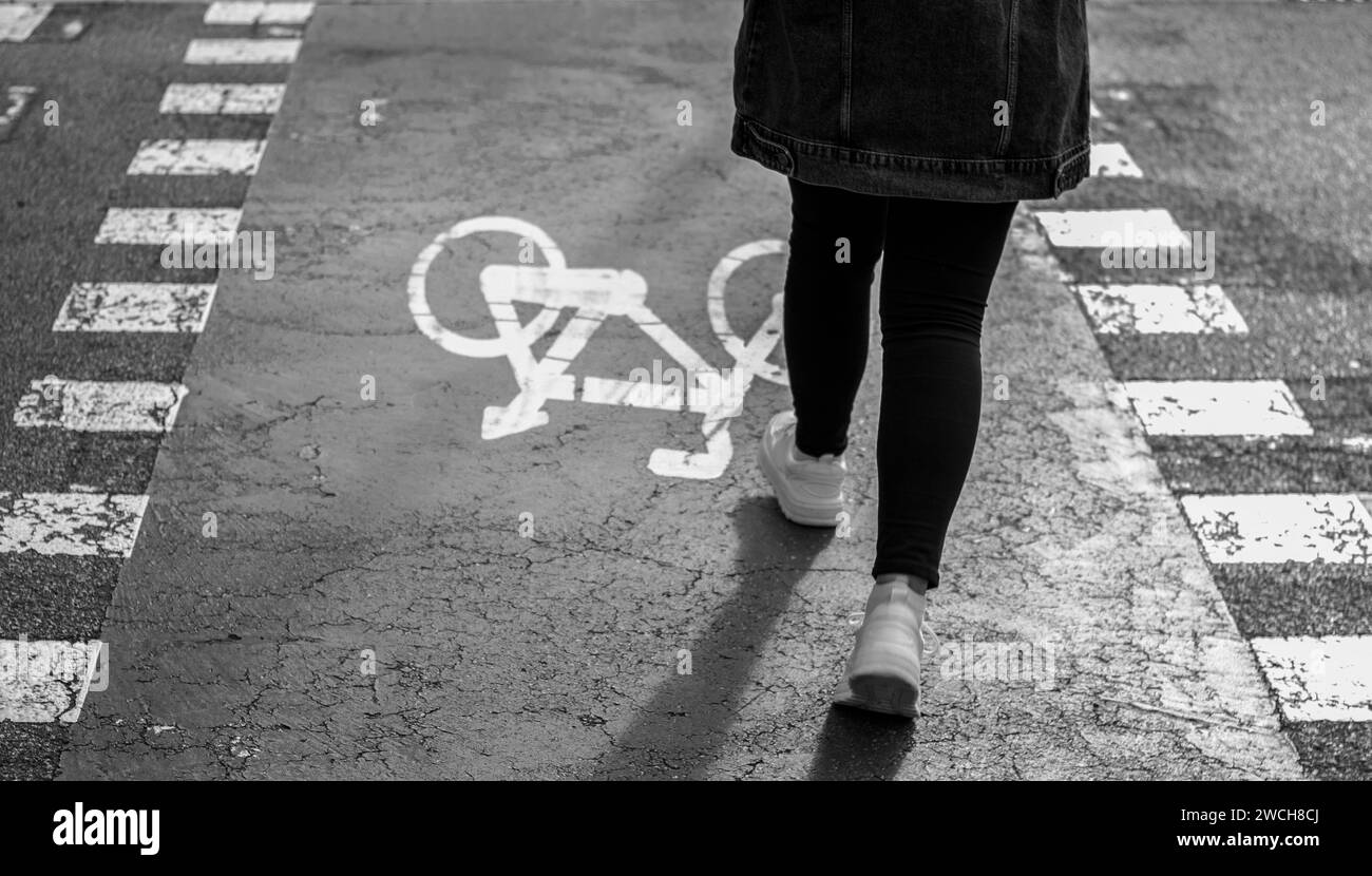 Das Mädchen in weißen Sneakers mit einer Jeansjacke geht zu Fuß über einen Fahrradweg. Fahrradschild auf Asphalt gemalt. Schwarzweißfoto. Stockfoto