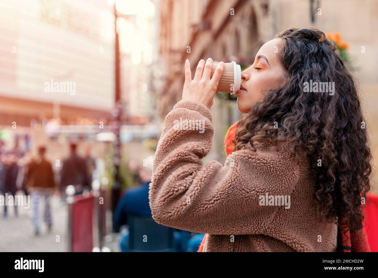 Eine fröhliche Frau, die eine Tasse Kaffee hält. Eine lächelnde, lockige Brünette in einem Pullover, die auf eine Straßenbahn wartet. Stockfoto