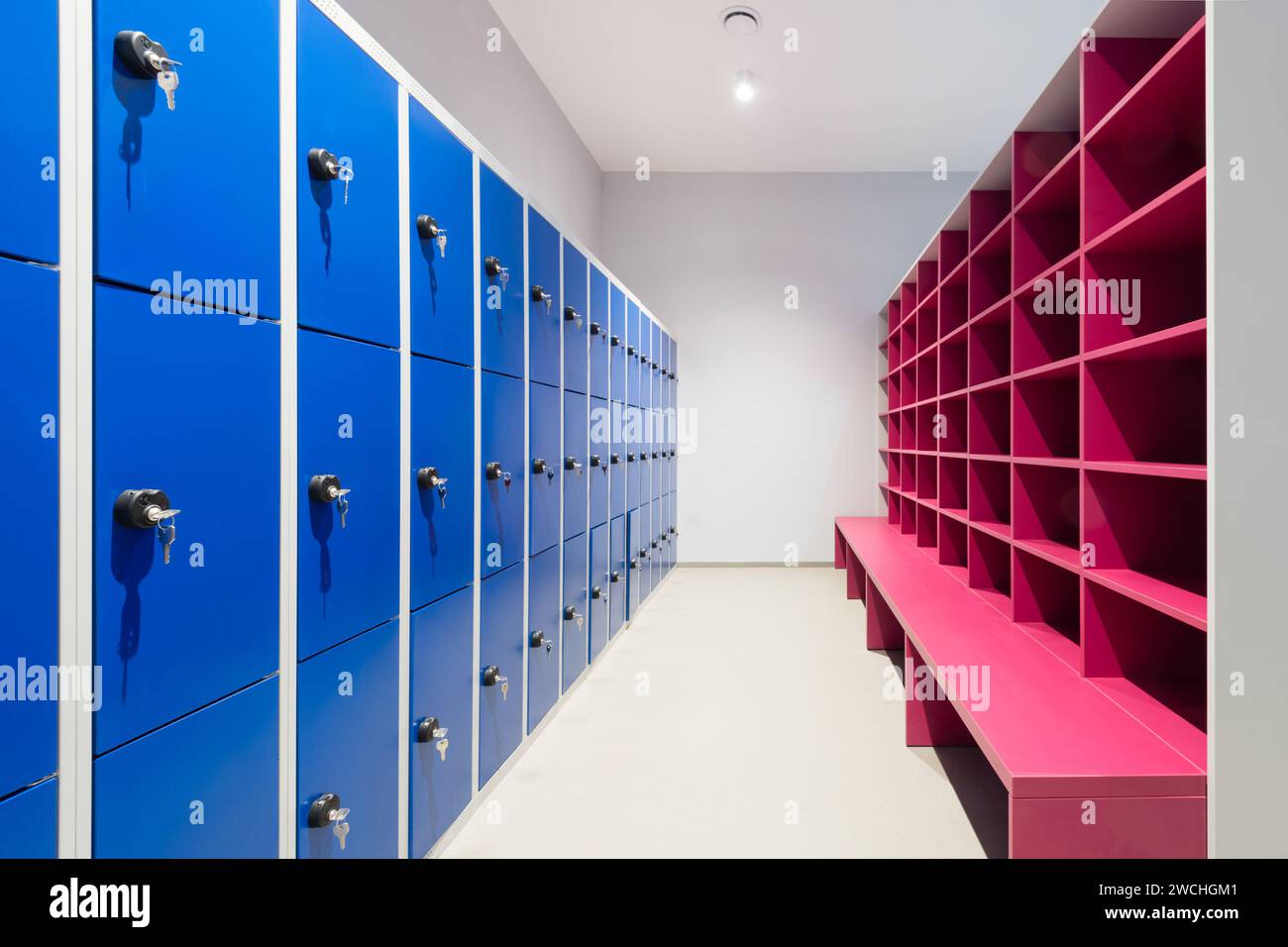 Eine Reihe blauer Schließfächer an einer Wand mit Schlüssel und rosa Schrank mit leeren Regalen und Bank Stockfoto