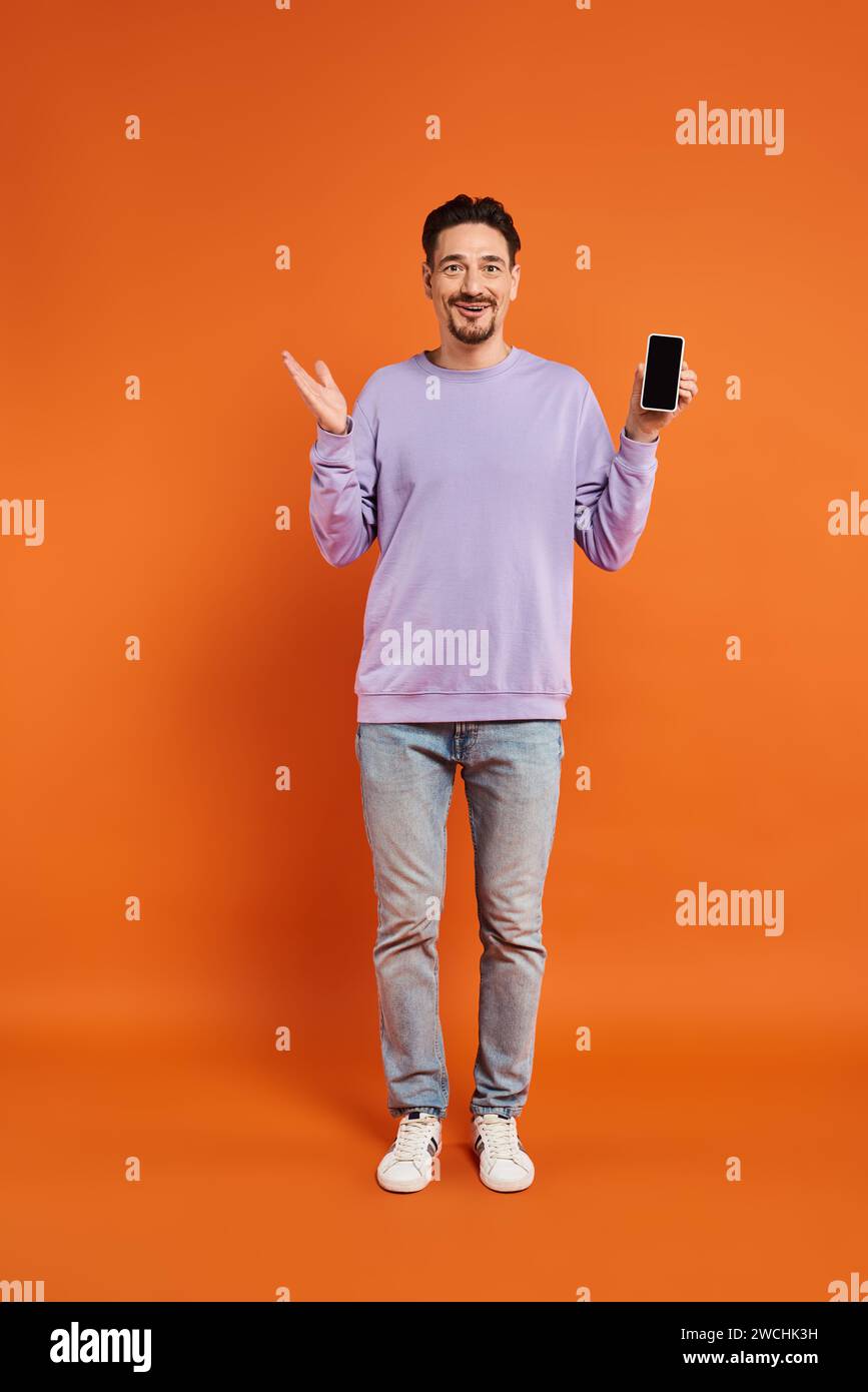 Staunender Mann in Brille und lila Pullover, der Smartphone mit leerem Bildschirm auf orangem Hintergrund hält Stockfoto