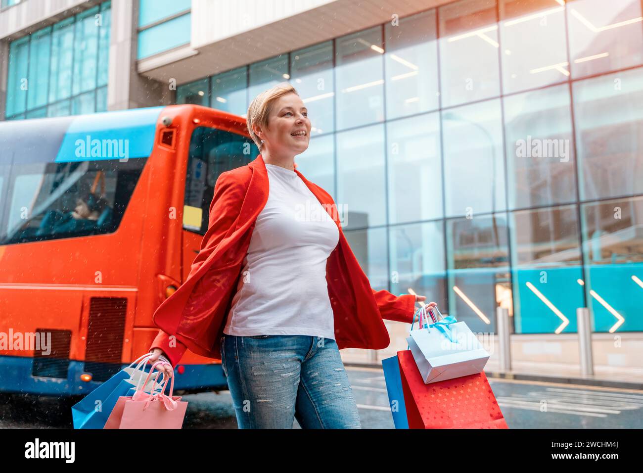 Glückliche Frau in roter Jacke mit bunten Taschen, die Spaß beim Einkaufen in der Stadt hat. Konsum, Verkauf, Einkäufe, Einkaufen, Lifestyle-Konzept Stockfoto