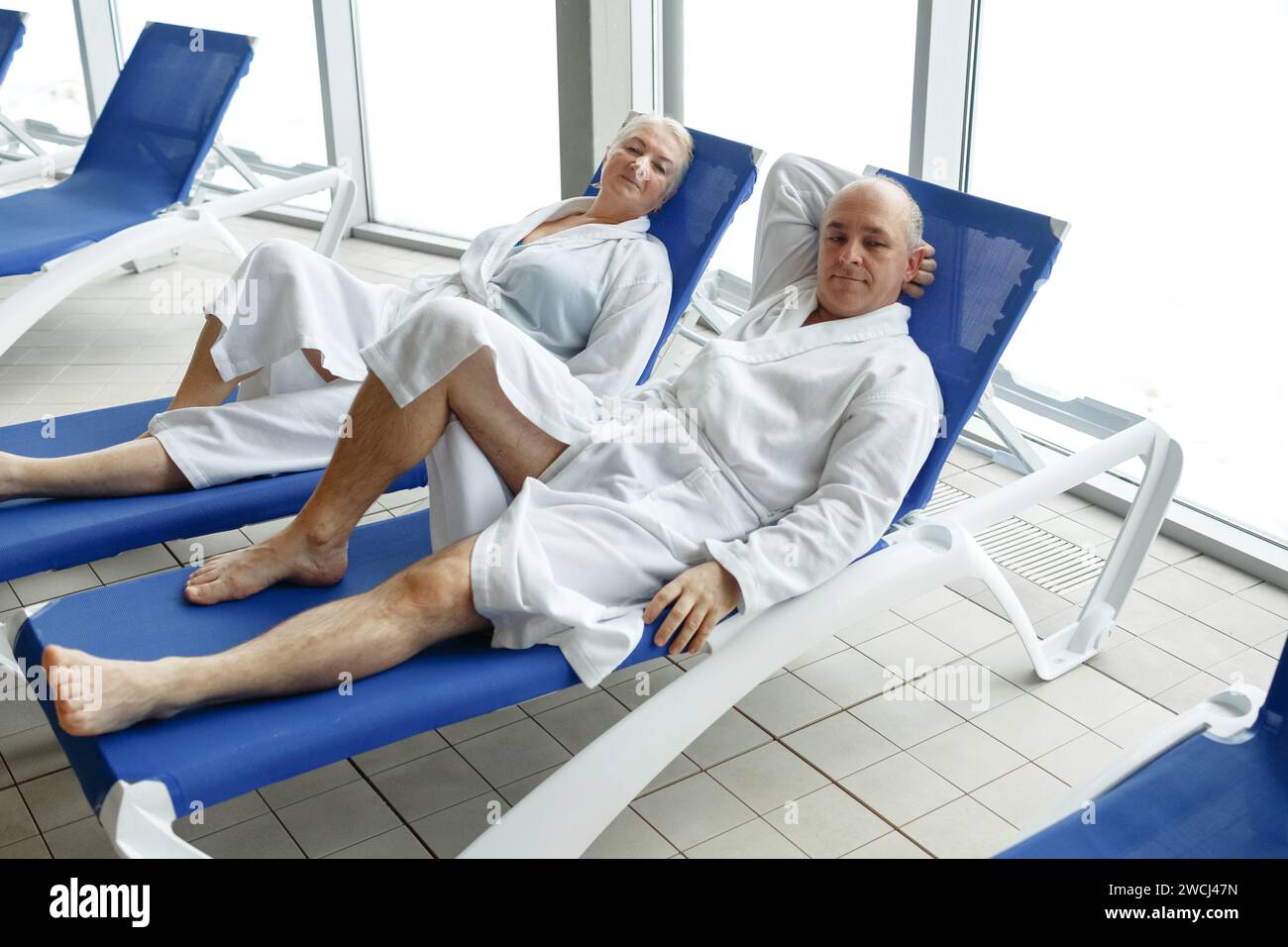 Eine ältere Frau mit einem Mann in Bademänteln auf einer Liege am Pool. Entspannen Sie sich im Spa-Center. Stockfoto