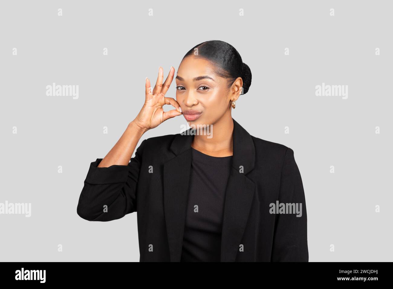 Eine fröhliche junge Frau in einem schwarzen Anzug macht ein verspieltes OK-Zeichen nahe ihrem Gesicht Stockfoto