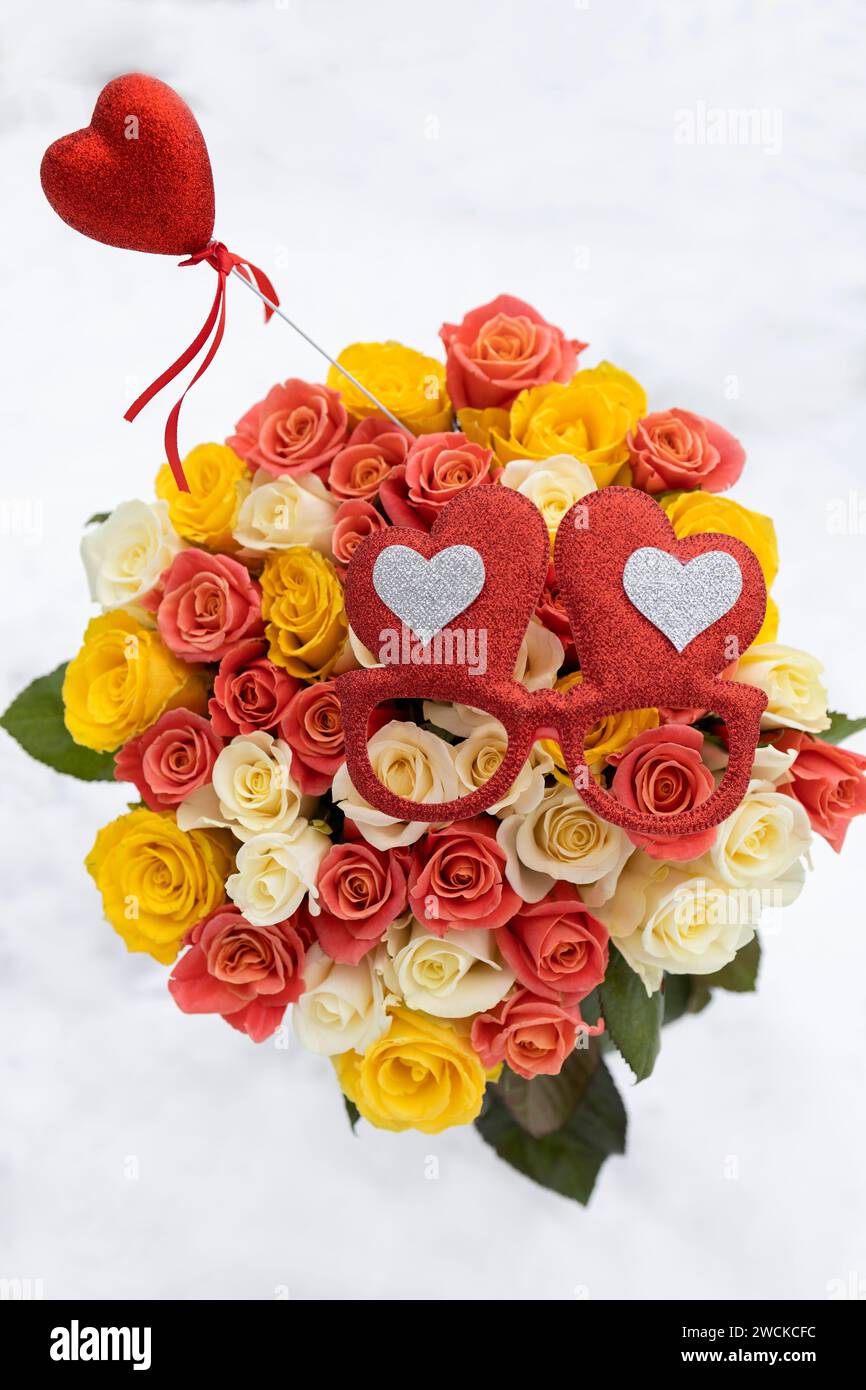 Buntes großes Blumenstrauß aus mehrfarbigen Rosen auf weißem Schneehintergrund. Rotes Souvenir-Herz und herzförmige Brille. Valentinstag. Stockfoto