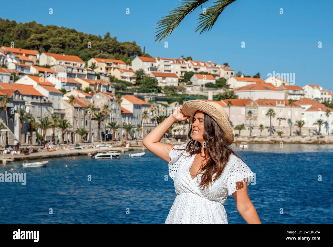 Eine elegante Frau steht anmutig am ruhigen Wasser und strahlt zeitlose Schönheit in ihrem fließenden weißen Kleid und ihrem stilvollen Strohhut aus Stockfoto