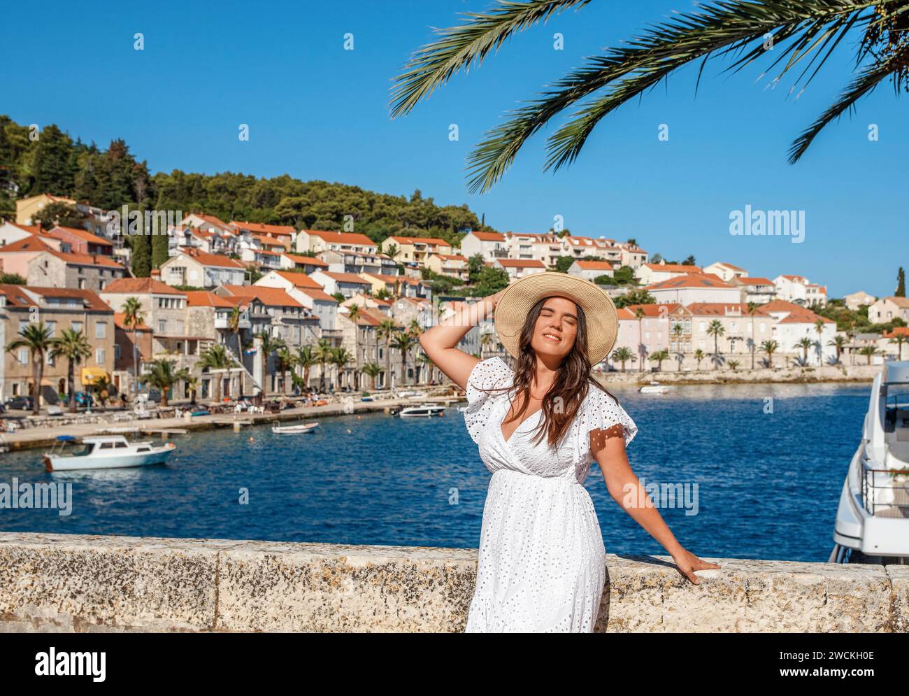 Eine atemberaubende junge Frau in einem eleganten weißen Kleid steht anmutig am malerischen Meer, mit einer bezaubernden Altstadt als Hintergrund Stockfoto