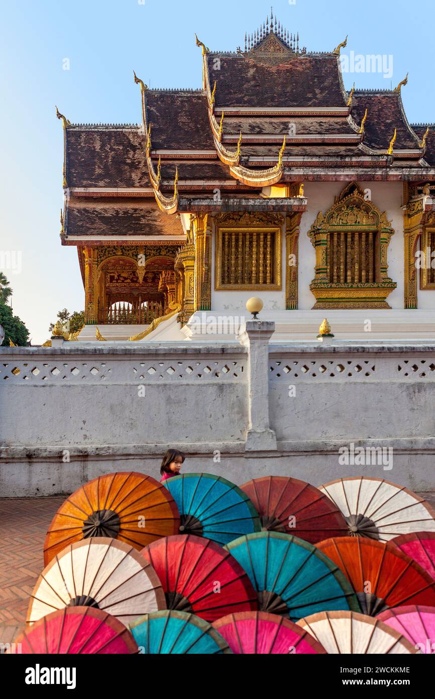 Kleines Mädchen, das hinter Schirmen vor dem Königspalasttempel (Haw Pha Bang) in Luang Prabang, Laos, läuft Stockfoto