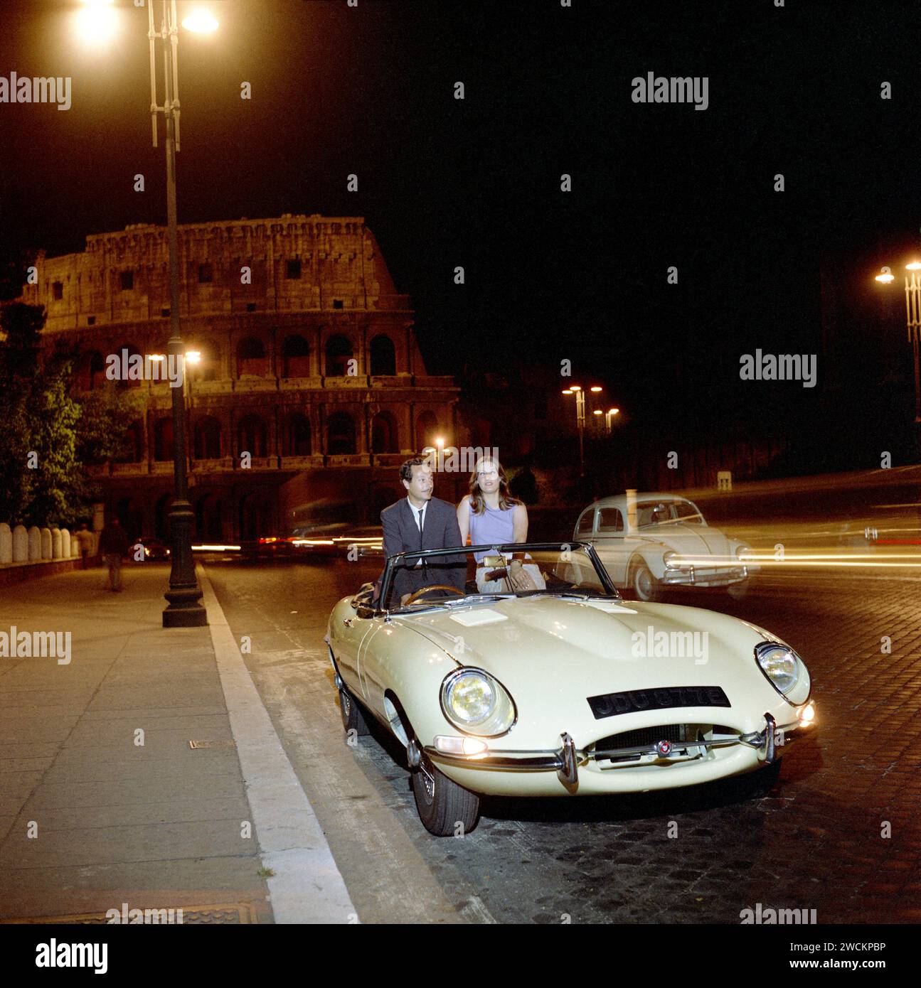 1967 Jaguar E-TYPE, Baureihe 1 1/2, in blassem Primrose Yellow, bei Nacht vor dem Kolosseum in Rom geparkt, mit einem romantischen jungen Paar. Stockfoto