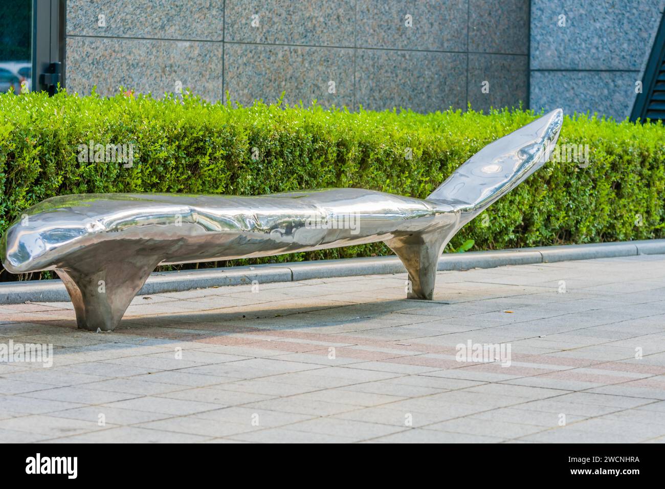 Große öffentliche Bank aus poliertem Chrom auf dem Bürgersteig vor grünem Heckenbusch Stockfoto