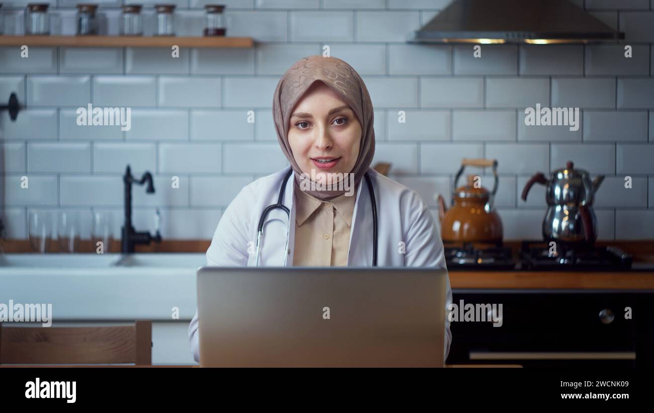 Die junge professionelle Ärztin im Kopftuch trägt einen weißen Mantel und spricht mit der Kamera, während sie in der Küche sitzt Stockfoto