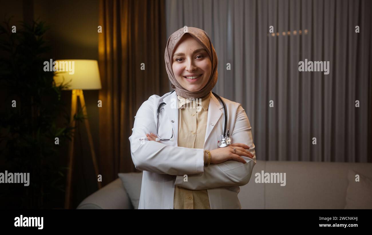 Porträt einer lächelnden Ärztin im Kopftuch trägt einen weißen Mantel und blickt nachts im Heimbüro in die Kamera Stockfoto
