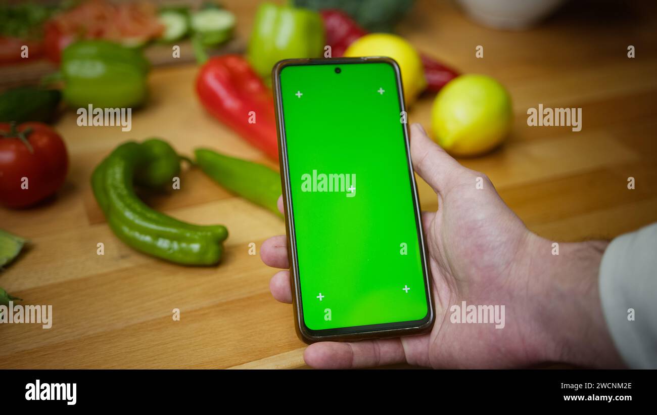 Mann, der ein Smartphone mit nachgeahmtem grünem Bildschirm-Chromakey-Display auf der Küchenarbeitsplatte mit grünem Gemüse benutzt Stockfoto