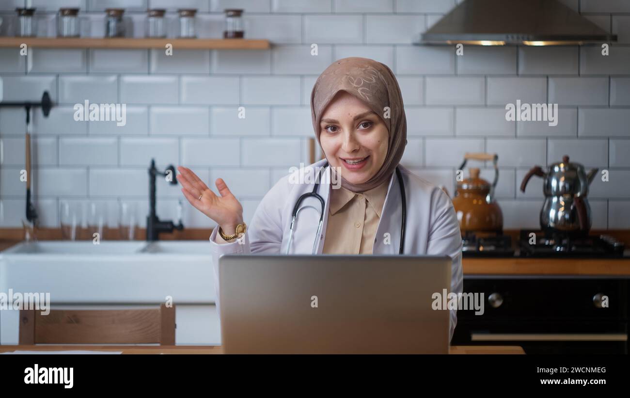 Die junge Ärztin im Kopftuch trägt einen weißen Mantel und spricht mit der Kamera, während sie in der Küche sitzt Stockfoto