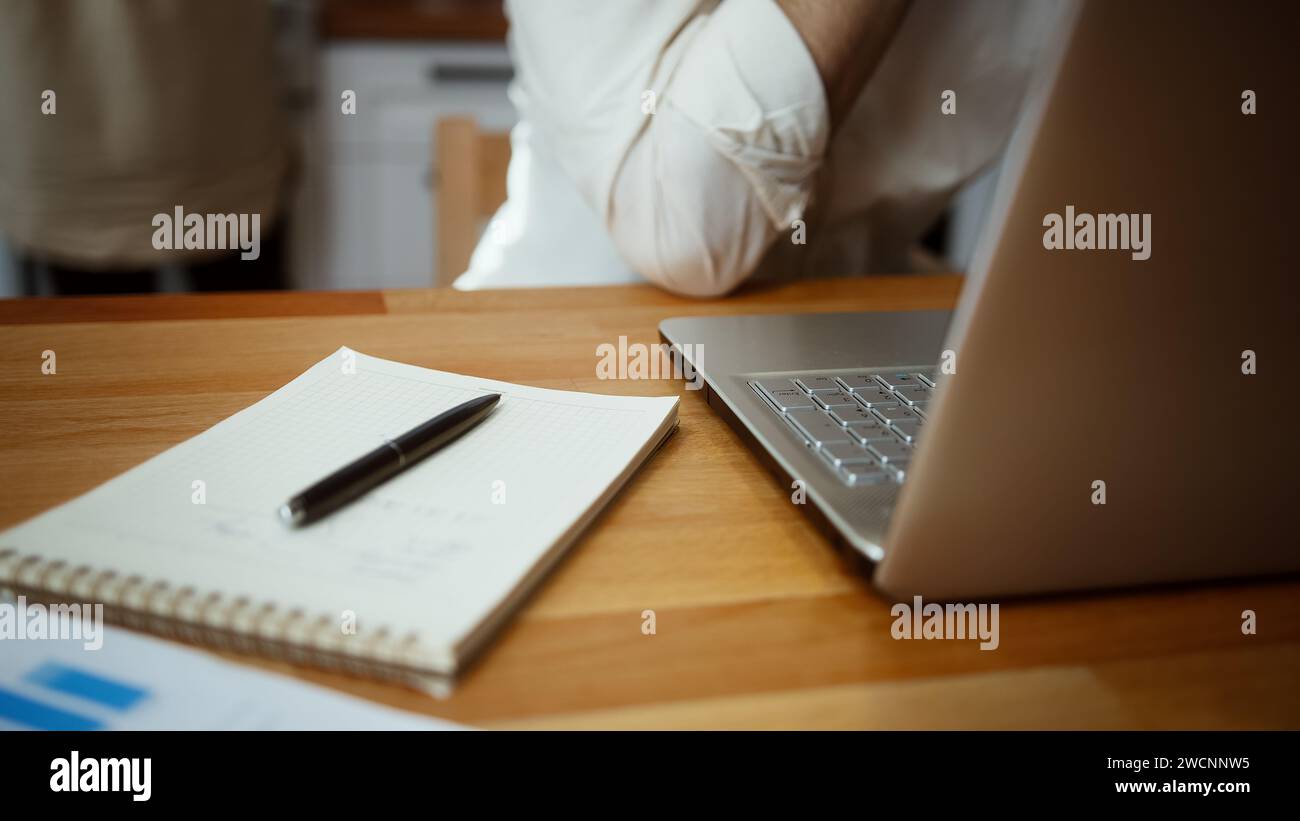 Mann, der online budgetiert, bezahlt Rechnungen und Finanzen, Hypothekenzahlungen mit Laptop-Computer, während seine Frau in der Küche arbeitet Stockfoto