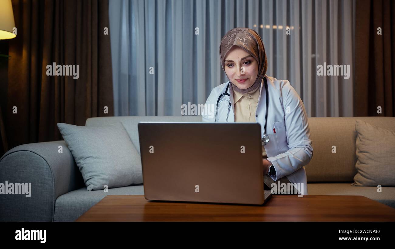 Die Ärztin im Kopftuch trägt einen weißen Mantel und spricht mit dem Patienten über die virtuelle Chat-Computer-App, während sie auf dem Sofa sitzt Stockfoto