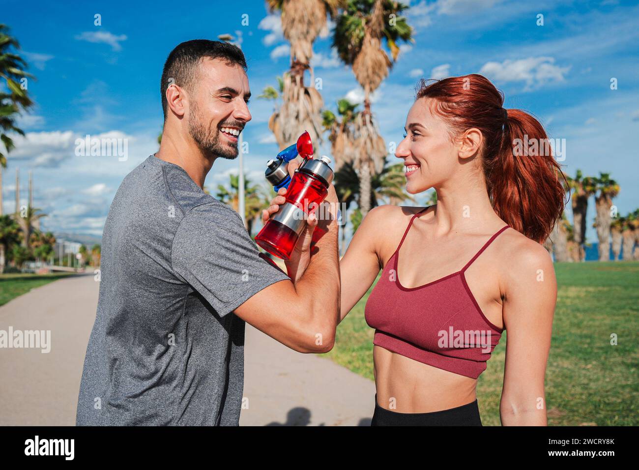 Sportpaare beim Toasten und Trinken von Wasser, während sie sich nach dem Laufen oder Workout ausruhen. Trainer Mann und starke Frau, lächelnd und klirrend ergänzen Flaschen, um eine Pause zu haben und sich während des Trainings zu erfrischen. Hochwertige Fotos Stockfoto