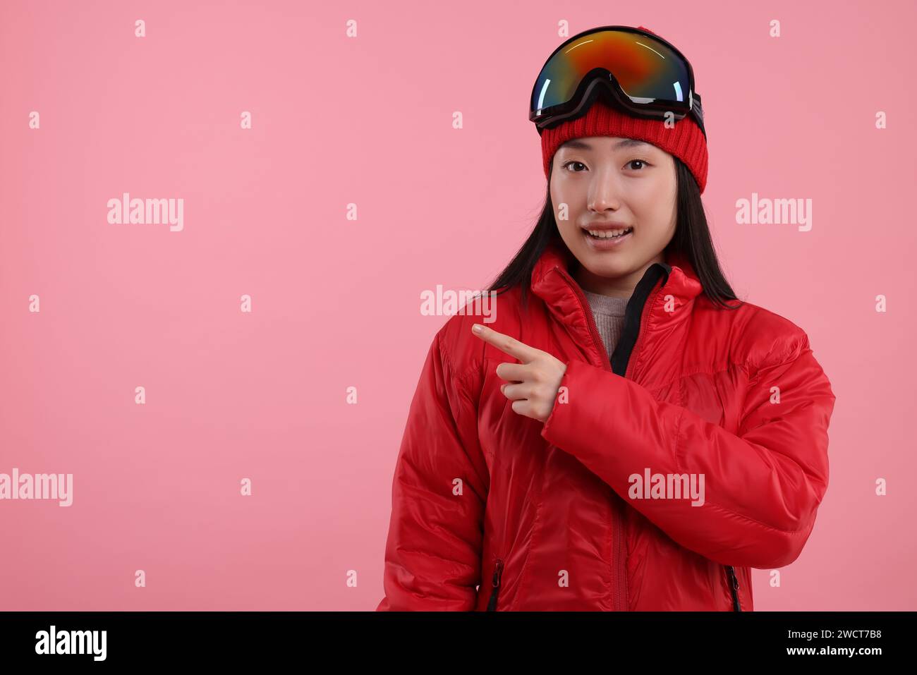 Glückliche Frau in Wintersportbekleidung, die auf etwas auf rosa Hintergrund zeigt. Leerzeichen für Text Stockfoto