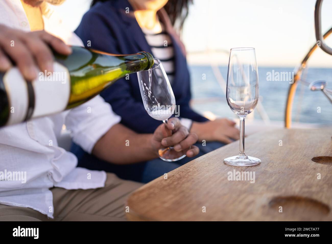 Ein Paar, das an einem Holztisch sitzt und Champagner in die Weingläser gießt, während es die Zeit auf der Yacht genießt Stockfoto